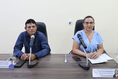 A imagem mostra vereador Benito do Estrela e a vereadora Bia do assentamento sentados lado a lado atrás de uma longa mesa de madeira marrom, olhando para a câmera. O ambiente é bem iluminado com uma parede branca lisa ao fundo. À esquerda, está o vereador Benito do Estrela de pele morena e cabelos curtos e escuros, vestindo uma camisa jeans azul escura de manga longa. Ele está com as mãos entrelaçadas sobre a mesa. À sua frente, há um microfone de mesa, um copo com água, um celular e uma placa de identificação. À direita, vereadora Bia do assentamento uma mulher de pele clara, cabelos loiros presos e usando óculos de grau. Ela veste uma blusa azul claro de manga curta com decote em V. Suas mãos estão apoiadas sobre algumas folhas de papel branco. À sua frente, também há um microfone, um celular e sua placa de identificação.