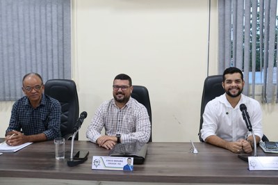 A imagem mostra três vereadores sentados atrás de uma mesa de madeira. O fundo apresenta uma parede branca e, à direita, uma janela coberta por persianas verticais cinzas. O vereador Gilmar do Detran à esquerda é mais velho, cor morena, usa óculos e uma camisa xadrez de manga longa em tons escuros. Ele olha ligeiramente para baixo, segurando uma caneta sobre papéis. O vereador Chico Moura ao centro, que sorri para a câmera, tem cabelos curtos, usa óculos, barba rala e uma camisa xadrez de manga longa em tons claros (branco e vinho). Ele está com as mãos entrelaçadas sobre a mesa, próximo a uma pasta preta e sua placa de identificação. O vereador Vitor Braz à direita é jovem, tem cabelo escuro penteado para trás, barba cheia e veste uma camisa social branca. Ele também sorri para a câmera com as mãos entrelaçadas. Há microfones de mesa distribuídos entre eles.